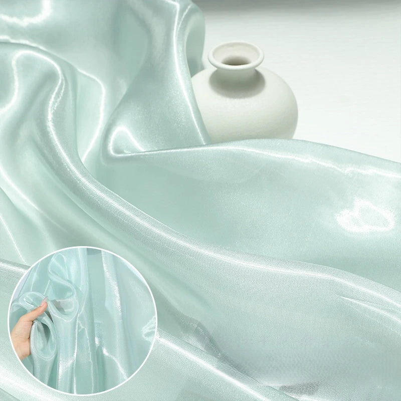 A close-up of Longan Craft's Shiny Liquid Organza Fabric By The Yard in light blue, with glossy finish, draped beside a white vase; inset shows a hand touching the sheer luxury gown material.
