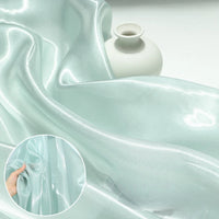 A close-up of Longan Craft's Shiny Liquid Organza Fabric By The Yard in light blue, with glossy finish, draped beside a white vase; inset shows a hand touching the sheer luxury gown material.