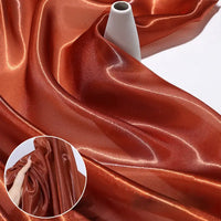 Longan Craft’s Shiny Liquid Organza Fabric By The Yard, with a colorful glossy finish, is elegantly draped around a white vase. An inset reveals a hand gathering the luxurious organza textile, perfect for luxury gown creations.