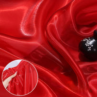 Shiny Liquid Organza Fabric by the yard from Longan Craft, with a colorful glossy finish, is elegantly draped and adorned with a black ceramic ornament. An inset highlights its luxurious texture as a hand gathers the vibrant organza textile.