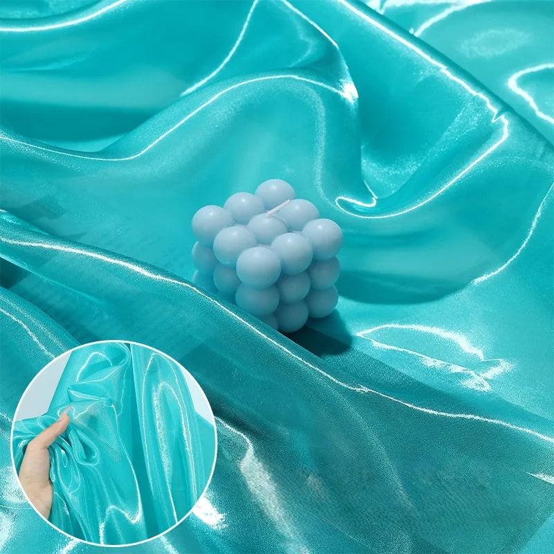 Shiny Liquid Organza Fabric By The Yard from Longan Craft features a colorful glossy finish in aqua blue, draped with a light blue bubble candle. Inset shows a hand crumpling this luxury gown material to display its elegant texture.