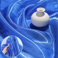 A small white ceramic vase sits on Longan Craft’s Shiny Liquid Organza Fabric By The Yard, a luxury organza textile with a colorful glossy finish. An inset shows a hand gathering this elegant material, ideal for evening or bridal gowns.