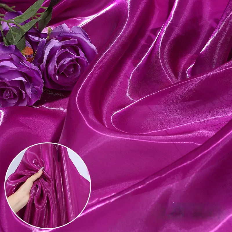 Shiny Liquid Organza Fabric by the Yard from Longan Craft features a vivid magenta, glossy finish with two artificial purple roses. An inset shows a hand gathering this luxury material—ideal for bridal gowns or evening wear.
