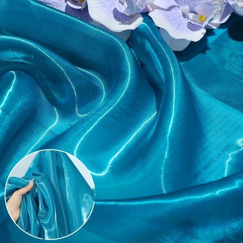Shiny Liquid Organza Fabric by Longan Craft features a colorful glossy finish that drapes smoothly with light reflections, ideal for luxury gowns. An inset shows a hand squeezing the fabric to display its rich texture. Available by the yard.