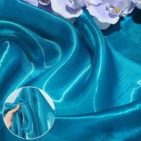 Shiny Liquid Organza Fabric by Longan Craft features a colorful glossy finish that drapes smoothly with light reflections, ideal for luxury gowns. An inset shows a hand squeezing the fabric to display its rich texture. Available by the yard.