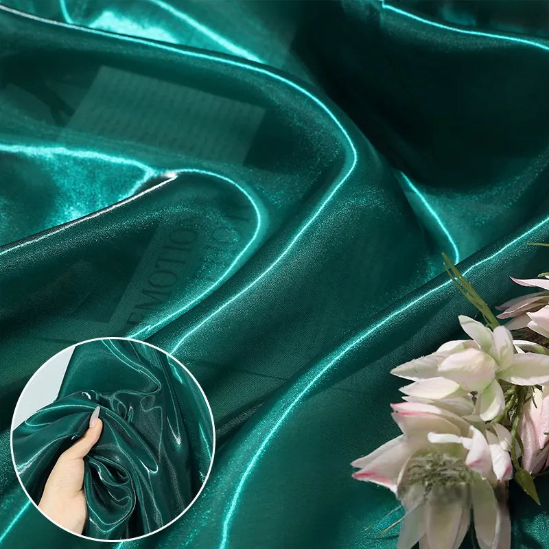 Shiny Liquid Organza Fabric by the yard from Longan Craft, featuring a colorful glossy finish and luxurious drape. Inset shows a hand holding the smooth organza; pink and white flowers accent the corner. Ideal for luxury gowns.
