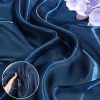 Longan Craft’s Shiny Liquid Organza Fabric By The Yard features a dark blue, glossy organza textile draped with light reflecting off its surface. Light purple flowers rest on top, and an inset shows a hand holding the sheer luxury material.