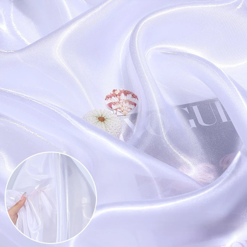 Shiny Liquid Organza Fabric by the yard from Longan Craft drapes smoothly in a sheer white with a colorful glossy finish; seashell and magazine details show its translucency—ideal for luxury bridal gowns.