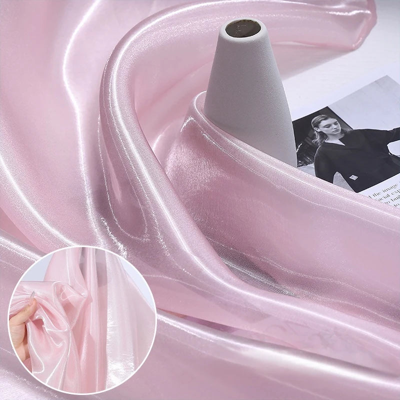 Shiny Liquid Organza Fabric by the yard from Longan Craft, shown in light pink draped on a white spool. This luxury, glossy finish organza textile is ideal for elegant bridal or evening gowns. Inset shows fingers holding the sheer material.