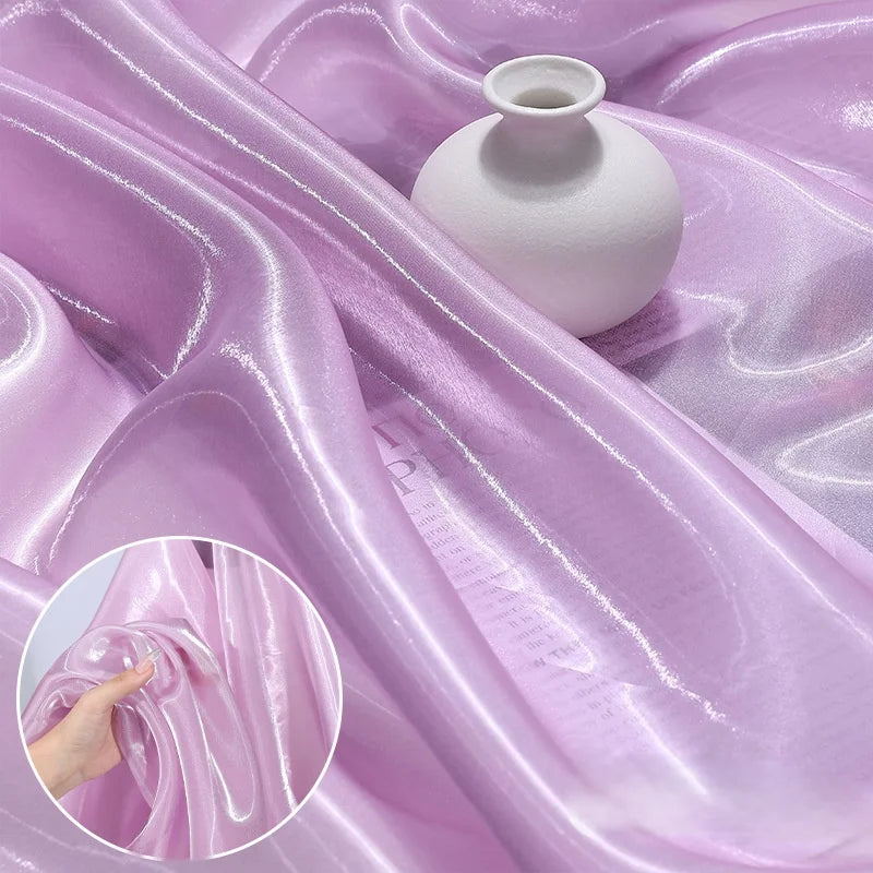 A white round vase sits on Shiny Liquid Organza Fabric by the Yard from Longan Craft, featuring a light purple, glossy finish. An inset displays a hand gathering the iridescent textile—ideal for luxury gowns or bridal wear.