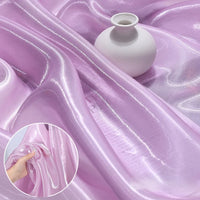 A white round vase sits on Shiny Liquid Organza Fabric by the Yard from Longan Craft, featuring a light purple, glossy finish. An inset displays a hand gathering the iridescent textile—ideal for luxury gowns or bridal wear.