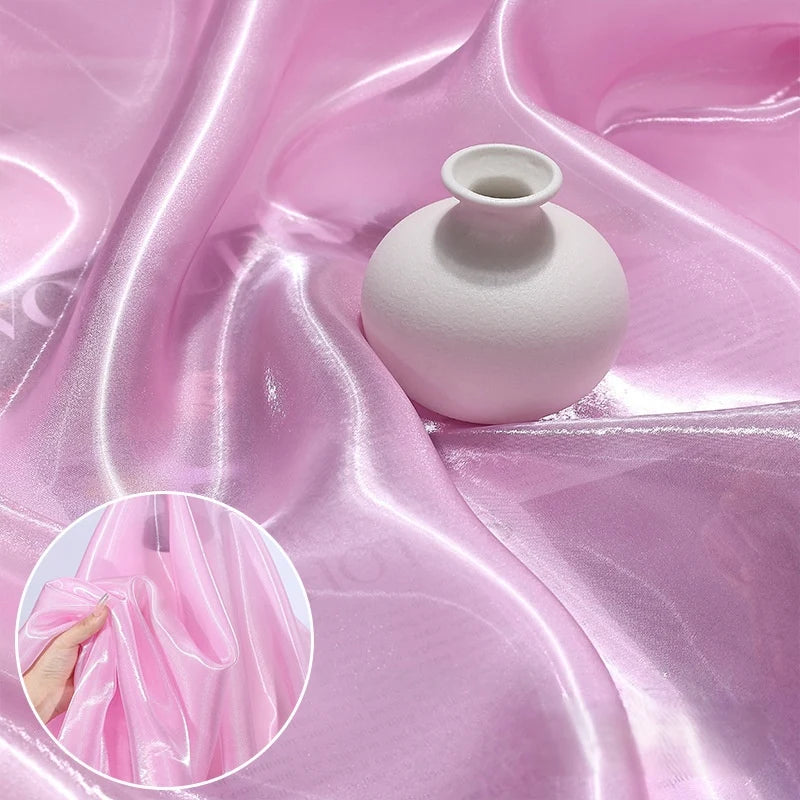 A small white ceramic vase rests on Longan Craft's Shiny Liquid Organza Fabric By The Yard—a luxury, glossy finish organza textile in light pink, ideal for gowns. Inset: a hand gently stretches the silky, luminous material to show its sheen.