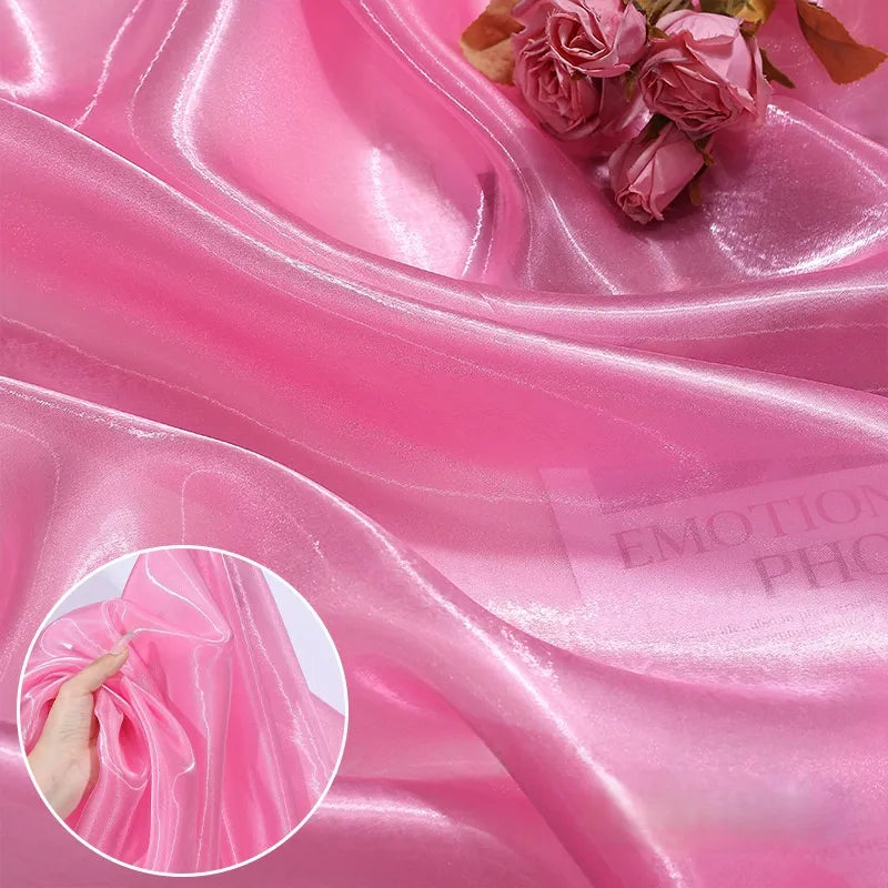 Longan Craft's Shiny Liquid Organza Fabric by the yard features a colorful, glossy finish. Perfect for luxury gowns or evening wear, this silky organza drapes beautifully—shown here with pink roses and a hand gathering its smooth texture.