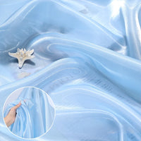 The Shiny Liquid Organza Fabric by Longan Craft features a light blue, glossy finish draped in soft folds with a starfish on top. An inset shows its translucent luxury texture and beautiful transparency for gown making.