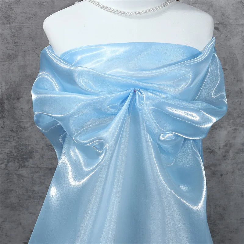A mannequin displays Longan Craft's Shiny Liquid Organza Fabric by the yard, a colorful glossy finish textile ideal for luxury gowns. Draped with a large bow and paired with a pearl necklace, it stands against a textured gray backdrop.