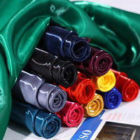 Various colors of Longan Craft's Shiny Liquid Satin Fabric By The Yard are rolled into rose shapes and displayed on a surface, with green satin draped above and a newspaper partially visible underneath.