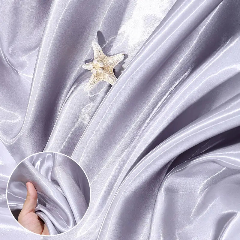 Shiny Liquid Satin Fabric by the Yard from Longan Craft is shown in soft light purple folds with a pale starfish on top. An inset displays a hand scrunching the glossy satin textile, perfect for formalwear or bridesmaid dresses.