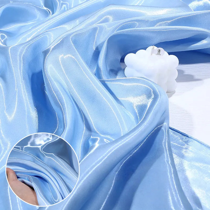 Shiny Liquid Satin Fabric by the Yard from Longan Craft in light blue drapes with soft folds, topped with a white bubble candle. Inset highlights a hand holding the smooth satin, perfect for bridesmaid dresses and formalwear.