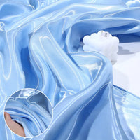 Shiny Liquid Satin Fabric by the Yard from Longan Craft in light blue drapes with soft folds, topped with a white bubble candle. Inset highlights a hand holding the smooth satin, perfect for bridesmaid dresses and formalwear.
