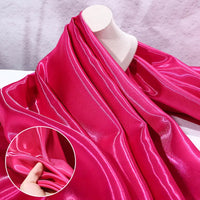 Longan Craft's Shiny Liquid Satin Fabric By The Yard in vibrant pink is displayed on a mannequin. A close-up shows a hand pinching the smooth, glossy textile, emphasizing its luxurious sheen—ideal for bridesmaid dresses and formalwear.