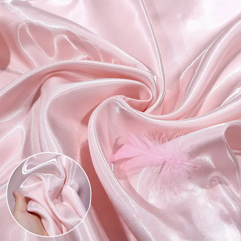 Longan Craft’s Shiny Liquid Satin Fabric By The Yard is shown in light pink, folded and gathered with a soft pink feather. An inset displays a hand holding the glossy satin, highlighting the material’s silky texture—ideal for formalwear.