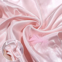 Longan Craft’s Shiny Liquid Satin Fabric By The Yard is shown in light pink, folded and gathered with a soft pink feather. An inset displays a hand holding the glossy satin, highlighting the material’s silky texture—ideal for formalwear.