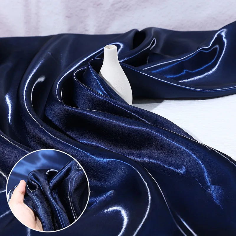 Longan Craft's Shiny Liquid Satin Fabric By The Yard in dark blue is draped around a small white vase. An inset shows the glossy, luxurious texture of this ideal satin material for bridesmaid dresses and formalwear as it's gently scrunched by hand.