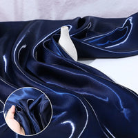 Longan Craft's Shiny Liquid Satin Fabric By The Yard in dark blue is draped around a small white vase. An inset shows the glossy, luxurious texture of this ideal satin material for bridesmaid dresses and formalwear as it's gently scrunched by hand.