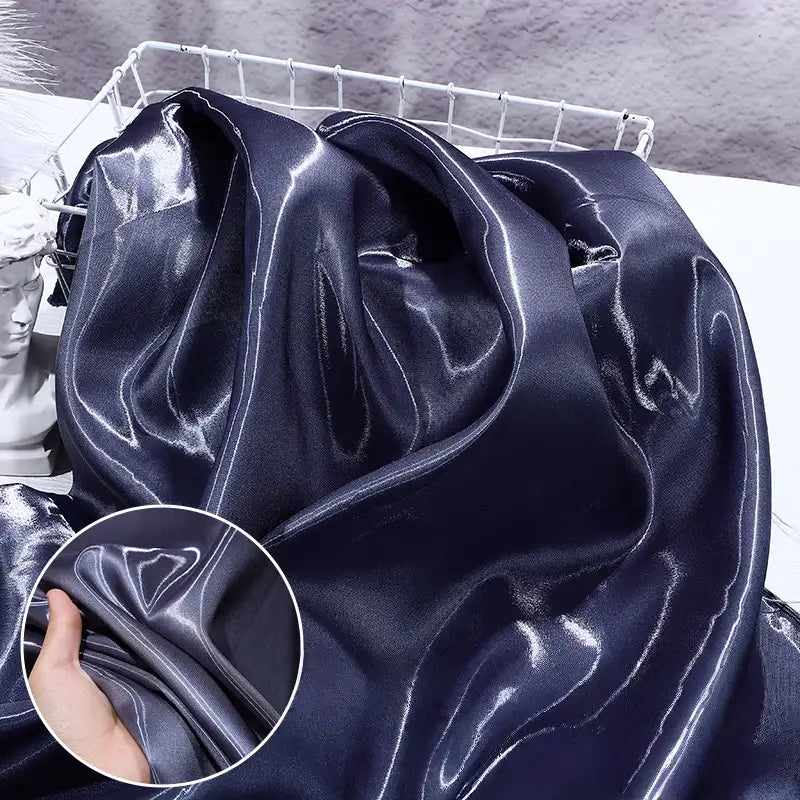 A smooth, shiny Longan Craft Liquid Satin Fabric in deep navy blue is draped over a surface, with a close-up of a hand holding the soft textile. Perfect for formalwear, sold by the yard.