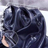 A smooth, shiny Longan Craft Liquid Satin Fabric in deep navy blue is draped over a surface, with a close-up of a hand holding the soft textile. Perfect for formalwear, sold by the yard.