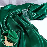 Shiny Liquid Satin Fabric by the yard from Longan Craft in emerald green is draped with a glass bottle, while an inset shows a hand pinching the smooth, glossy material—ideal for bridesmaid dresses and formalwear.