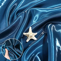 Longan Craft’s Shiny Liquid Satin Fabric by the yard, in bright blue with a glossy, reflective surface draped in folds and a white starfish on top. Inset: hand pinching this smooth satin—ideal for solid color bridesmaid dresses and formalwear.