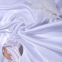 Longan Craft’s Shiny Liquid Satin Fabric By The Yard features smooth, shiny polyester satin ideal for bridesmaid dresses and formalwear. Image shows the luxurious white fabric gathered with a pearl-accented feather and a hand highlighting its soft texture.