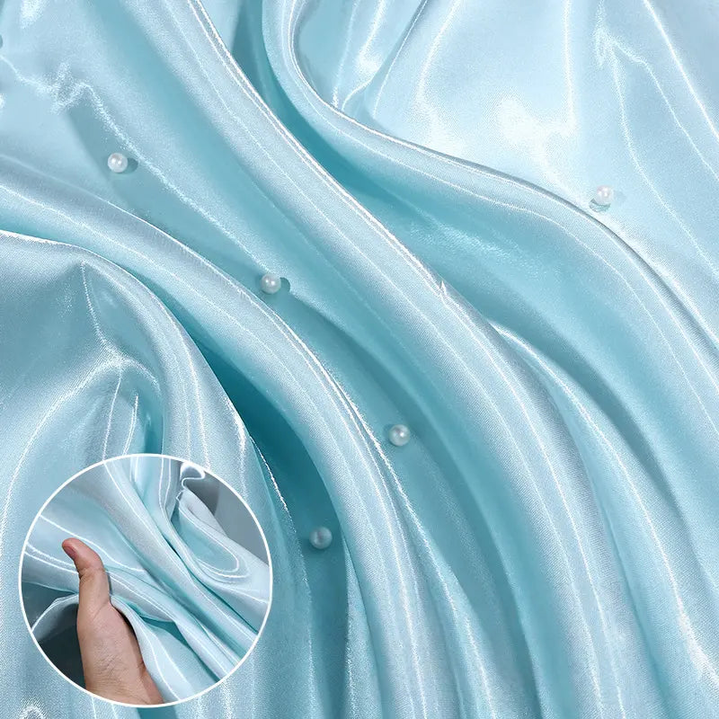 Close-up of Longan Craft’s Shiny Liquid Satin Fabric By The Yard in light blue with pearl accents. An inset displays a hand gathering the glossy polyester satin, highlighting its luxurious texture—ideal for formalwear and bridesmaid dresses.