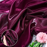 Shiny Liquid Satin Fabric by Longan Craft in deep magenta is draped beside a pink carnation. An inset shows a hand crumpling the glossy, soft satin, highlighting its sheen—perfect for bridesmaid dresses and formalwear.