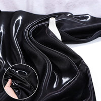 Longan Craft’s Shiny Liquid Satin Fabric By The Yard in deep black, ideal for formalwear and bridesmaid dresses, is draped over a white surface with a small vase. An inset highlights the material’s soft, glossy polyester texture.
