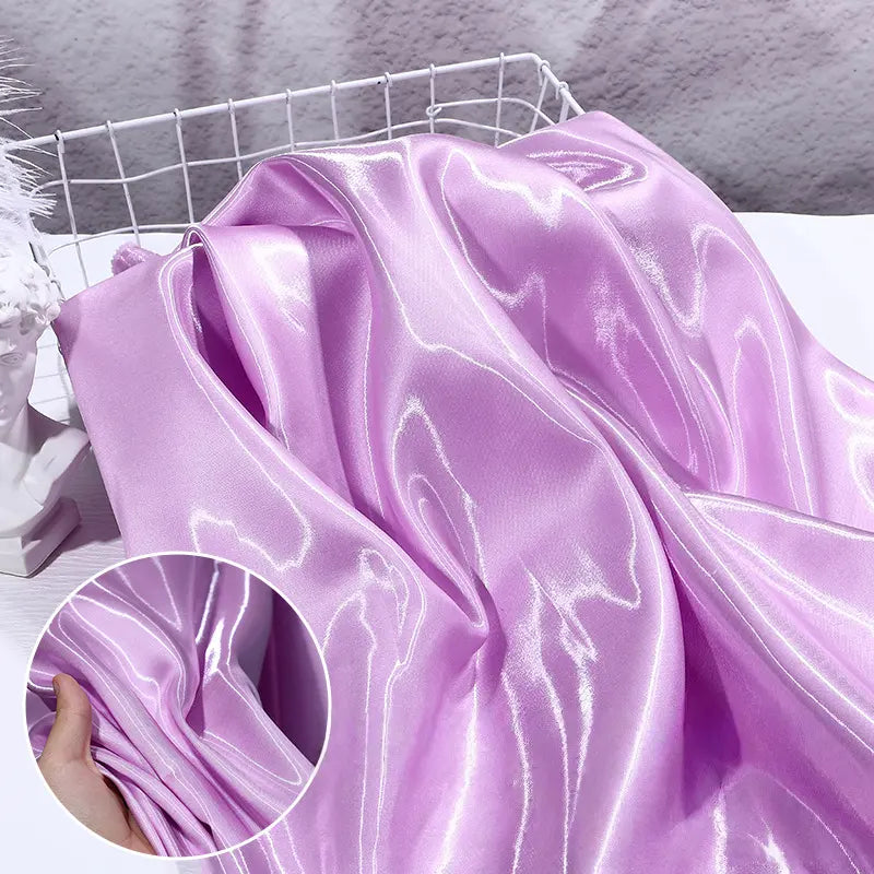A close-up of Longan Craft’s Shiny Liquid Satin Fabric By The Yard in light purple, highlighting its smooth, reflective texture. An inset shows a hand gathering the luxurious material, ideal for formalwear and bridesmaid dresses.
