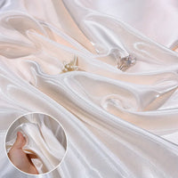 Shiny Liquid Satin Fabric by Longan Craft is elegantly draped in soft folds. Two delicate rings, including a butterfly design, rest atop the smooth textile. An inset highlights its silky texture as a hand gently pinches the fabric.
