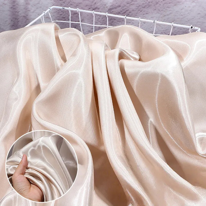 A close-up of Longan Craft’s Shiny Liquid Satin Fabric By The Yard, draped over a white wire basket, with an inset showing a hand gently scrunching the soft, glossy bridal satin—ideal for formalwear and bridesmaid dresses.