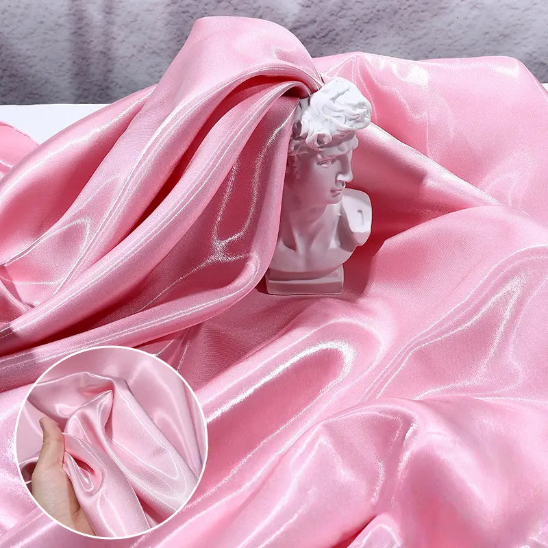 A shiny pink Longan Craft Liquid Satin Fabric By The Yard drapes around a small white bust. An inset features a hand bunching the smooth, glossy fabric to showcase its luxurious texture, perfect for bridesmaid dresses and formalwear.