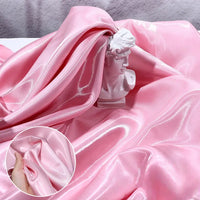 A shiny pink Longan Craft Liquid Satin Fabric By The Yard drapes around a small white bust. An inset features a hand bunching the smooth, glossy fabric to showcase its luxurious texture, perfect for bridesmaid dresses and formalwear.