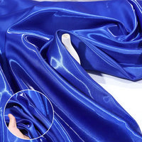 Shiny Liquid Satin Fabric by Longan Craft in royal blue is draped with smooth folds. An inset shows a hand feeling its soft, glossy texture and vibrant color—ideal for bridesmaid dresses and formalwear.