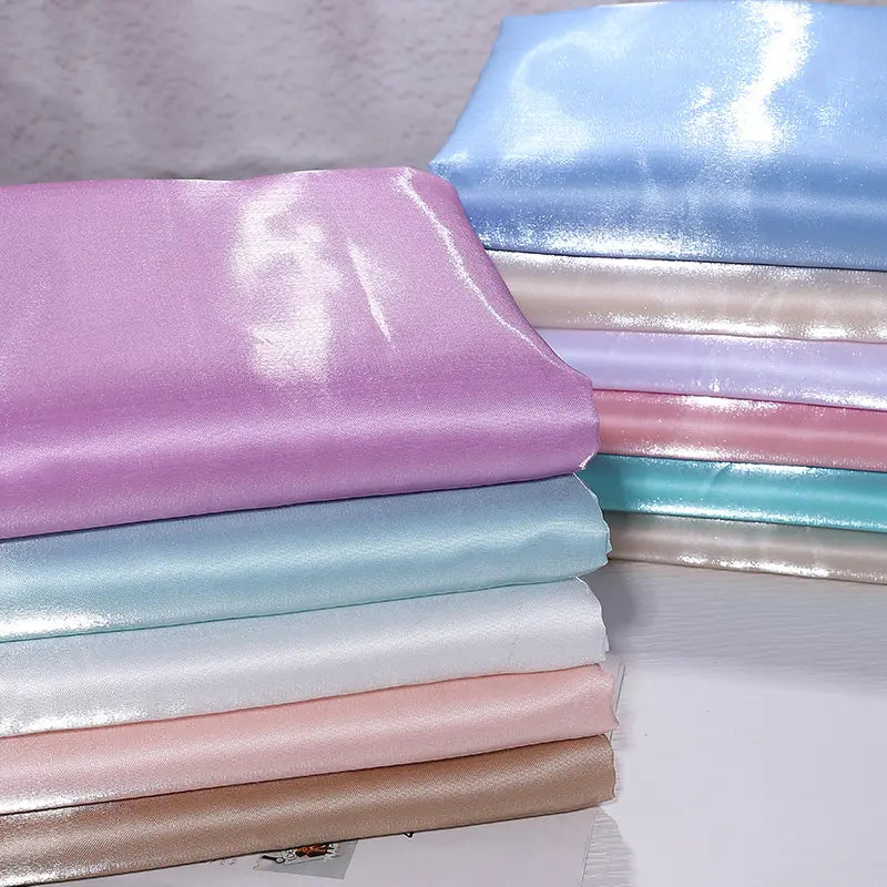 A stack of Longan Craft’s Shiny Liquid Satin Fabric By The Yard in purple, blue, white, pink, and beige is neatly folded on a textured white surface—ideal solid satin material for bridesmaid dresses and formalwear.