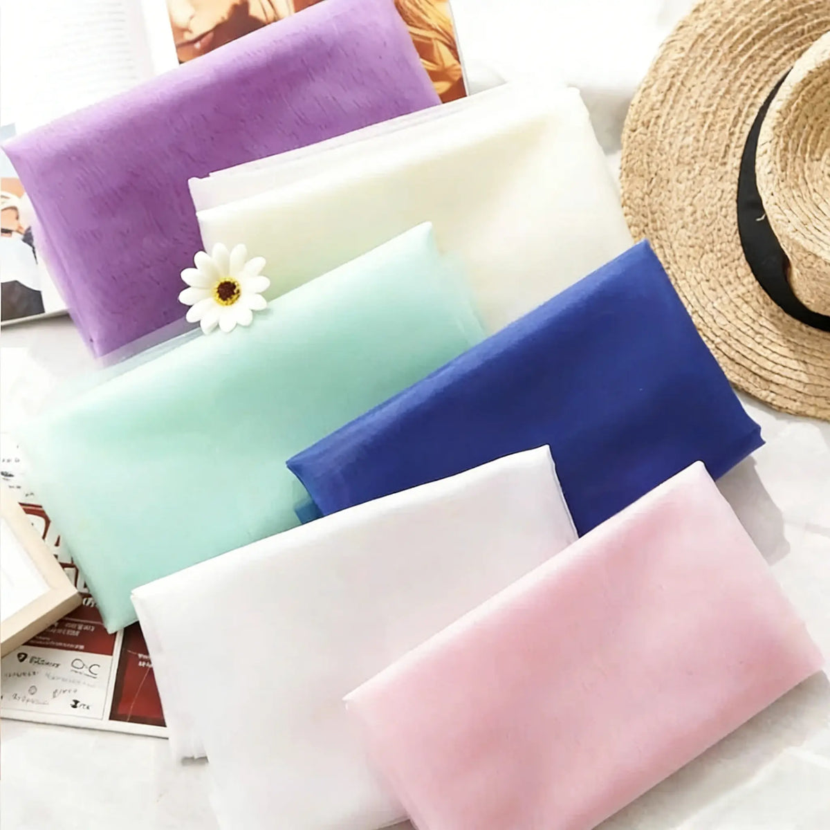 Premium Silky Polyester Organza Fabric by the yard from Longan Craft in pastel shades (purple, white, green, blue, pink) is arranged with a straw hat, open book, and flower—ideal inspiration for evening dresses or bridal gowns.