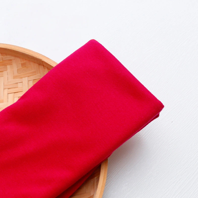 A piece of bright red Single Jersey Fabric by Longan Craft rests on a round bamboo tray against a white background. This colorful polyester jersey cloth is breathable, perfect for casual wear and available by the yard.