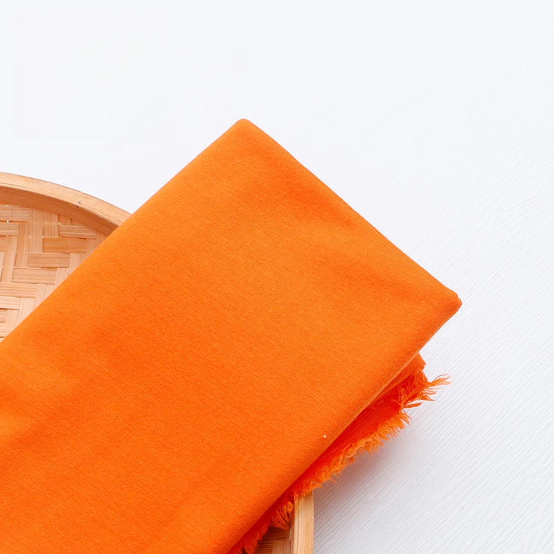 A piece of bright orange Single Jersey Fabric by the yard from Longan Craft, featuring colorful polyester jersey material for casual wear, is displayed with frayed edges on a round bamboo tray against a white background.