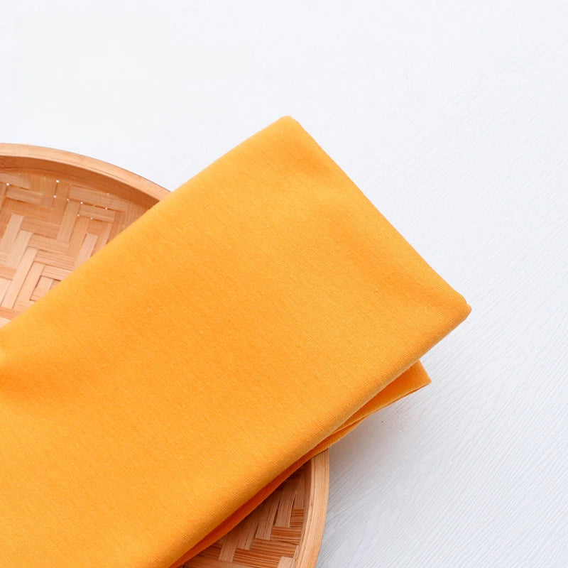 A vibrant yellow Single Jersey Fabric by Longan Craft is neatly folded on a round woven bamboo tray, displayed against a white background. Perfect colorful polyester jersey material for casual wear and DIY projects.