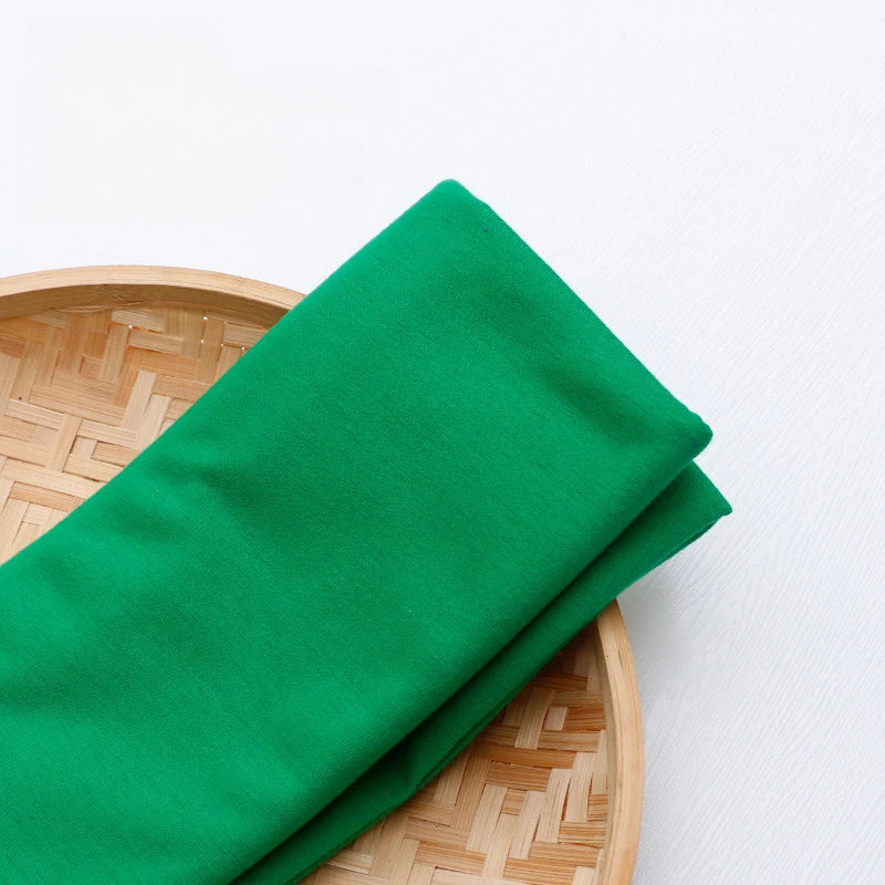 A vibrant green Single Jersey Fabric by Longan Craft, crafted from breathable polyester and perfect for casual wear, is neatly folded on a round bamboo tray atop a white surface.