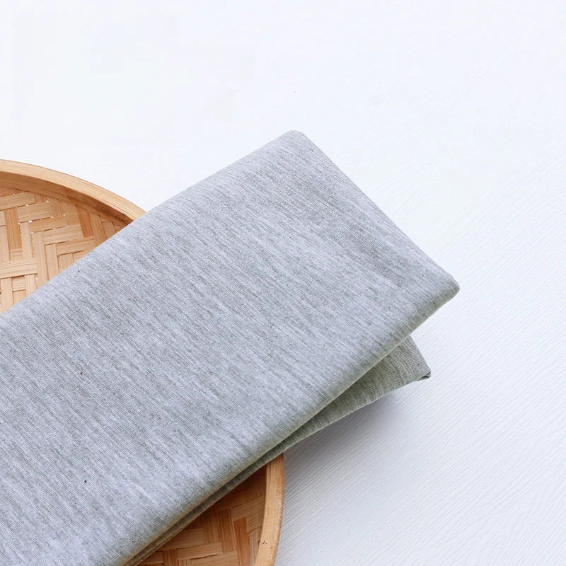 A piece of Longan Craft Single Jersey Fabric By The Yard in light gray rests on a woven bamboo tray, showcasing this colorful polyester jersey cloth ideal for casual wear.