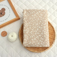 A folded piece of “Small Floral Print Double Gauze Fabric By The Yard” from Longan Craft, featuring a botanical print on gentle cotton crepe gauze, rests on a round woven mat beside a wooden button, a white yarn ball, and a framed bear illustration atop a quilted white surface.